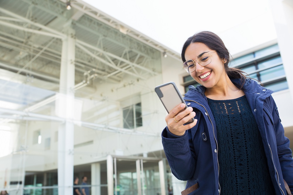 Joyful carefree office girl watching a 360 virtual tour on phone. Young woman in overcoat standing near building glass wall and laughing at phone screen. Wireless connection concept