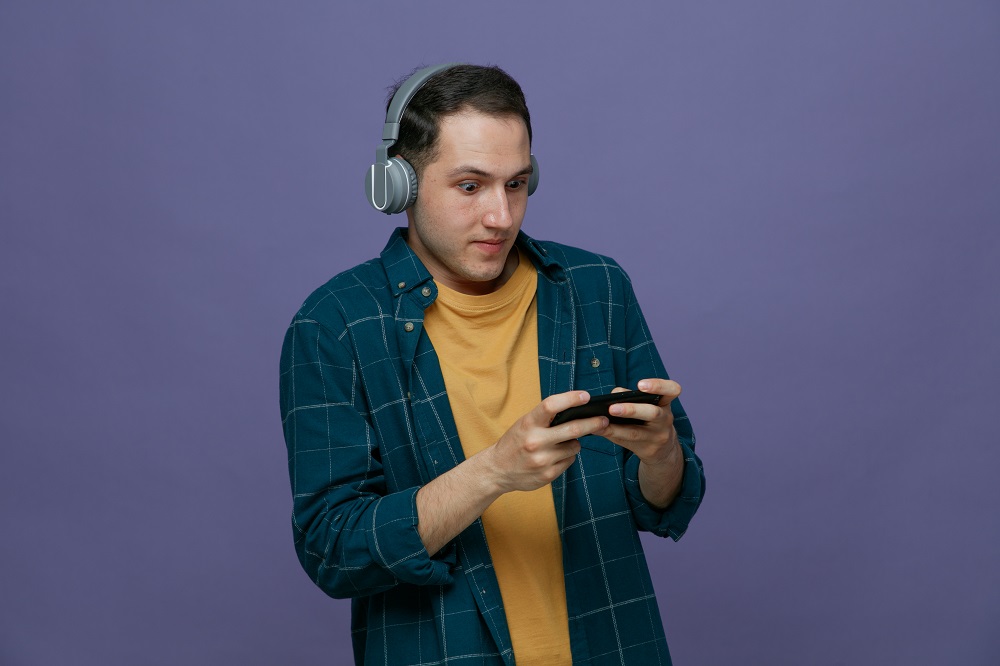 impressed young male student wearing headphones playing interactive games on mobile phone