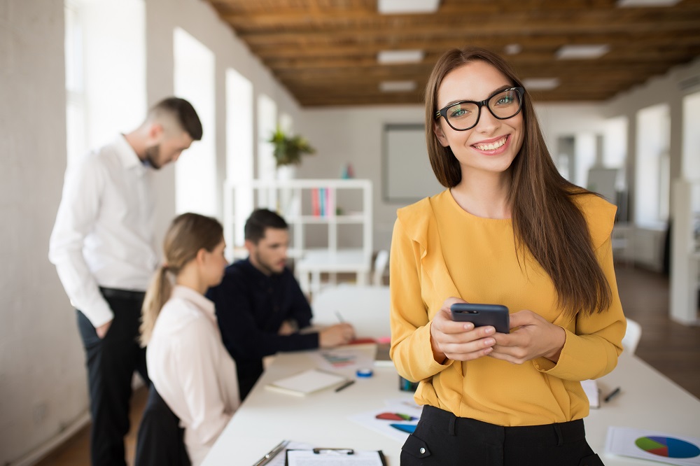 Woman in eyeglasses happily looking in camera holding cellphone in hands in office with colleagues on background uses a mobile app